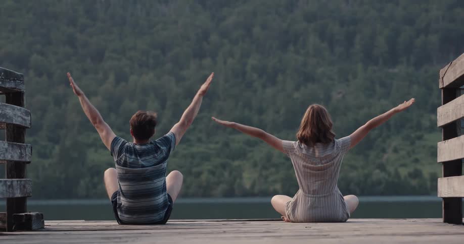 A man and woman sit cross legged on a dock stretching while facing the lake