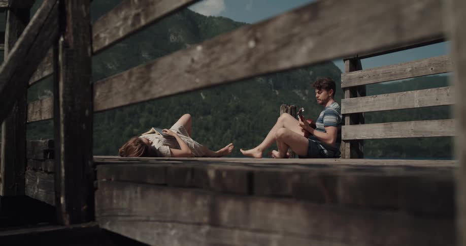 Man with shorts and t shirt plays guitar next to woman lying down on pier against the high mountains