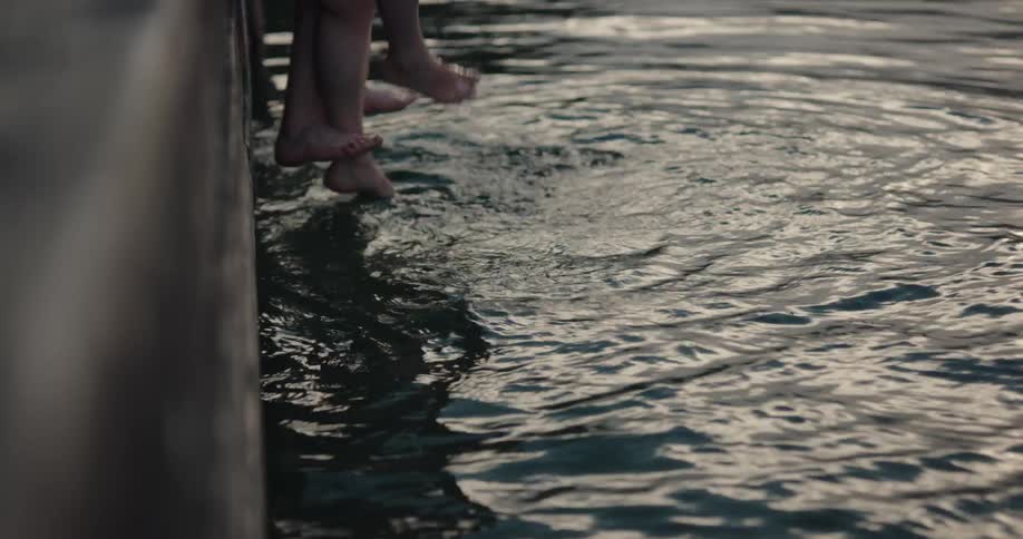 A man and woman splash their feet in water as they sit on a pier over a lake