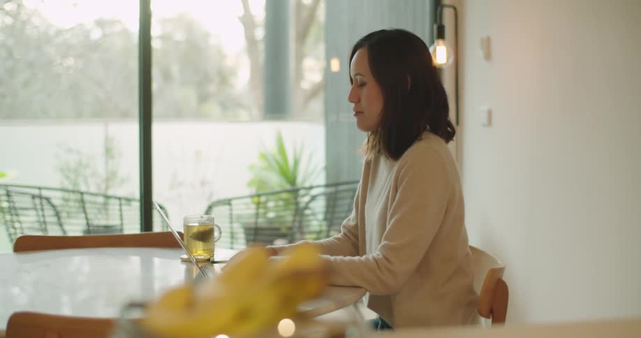 Woman sitting at table looks outdoors briefly before typing on laptop