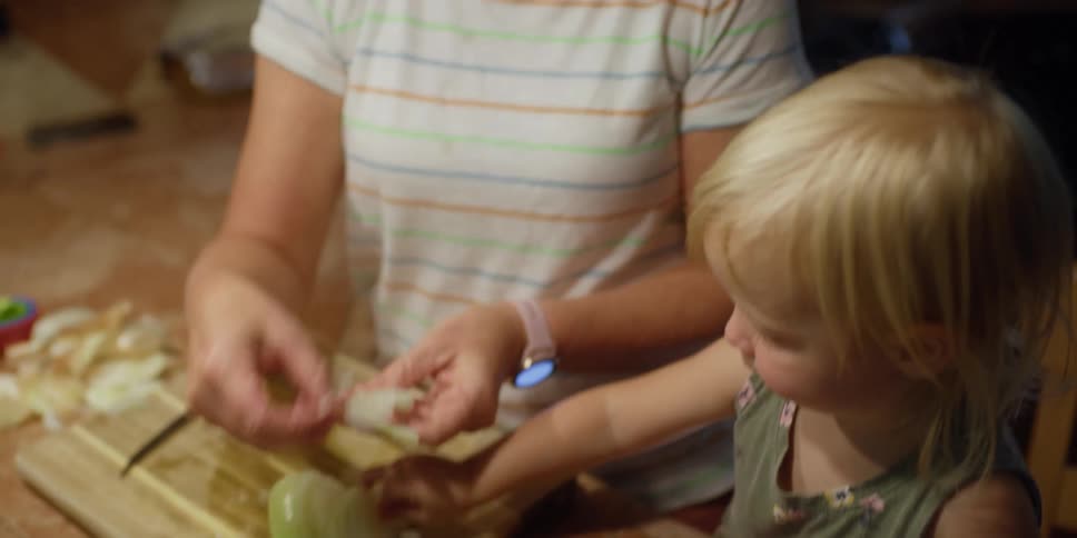 A young toddler helps a woman place sliced onion in a pan