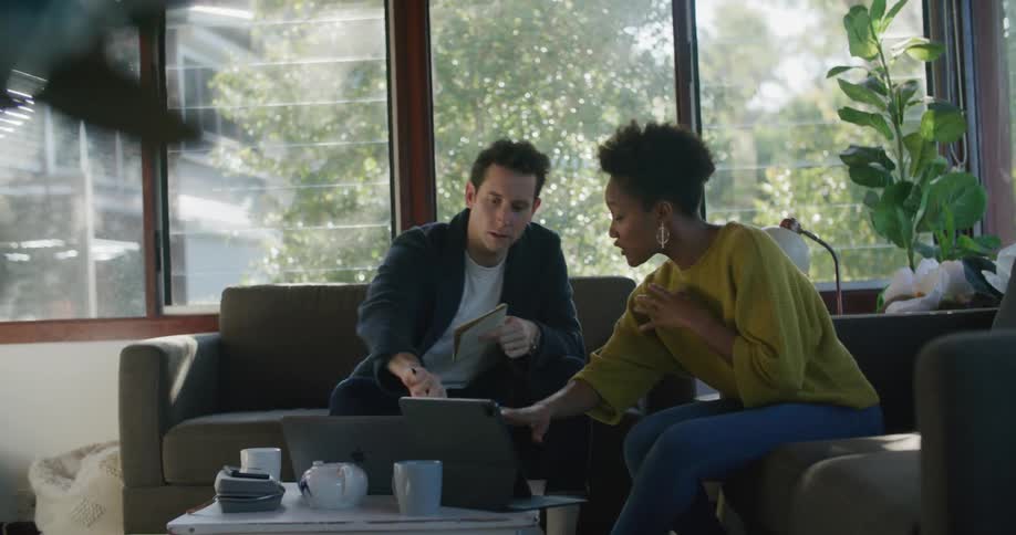 Man and woman sit in living room near windows consulting over laptop and tablet