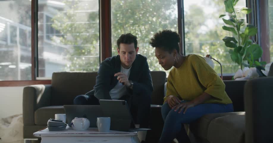 A woman talks to a man sitting next to her on a sofa while gesturing at the screen of her laptop