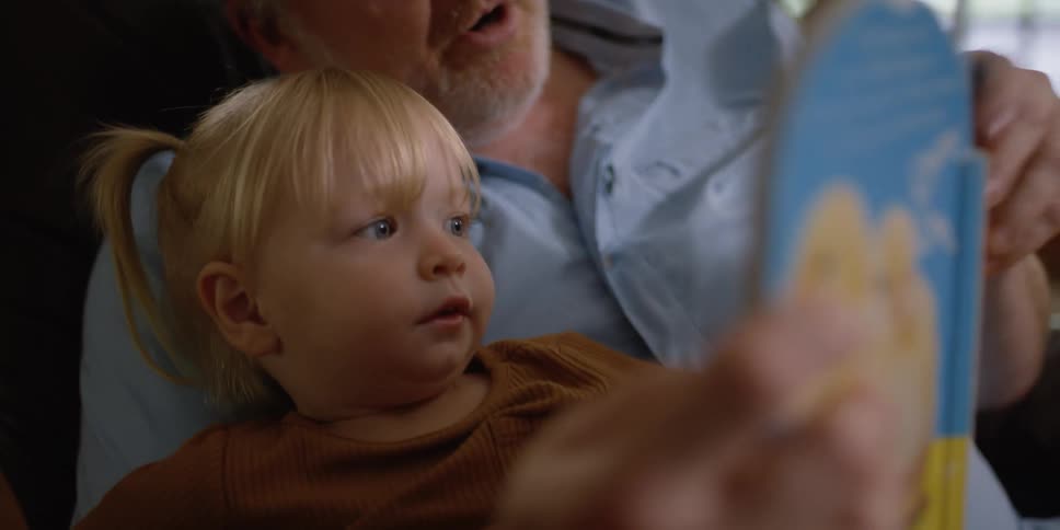 Grandfather with a beard and blue shirt holds granddaughter and shows her a story book