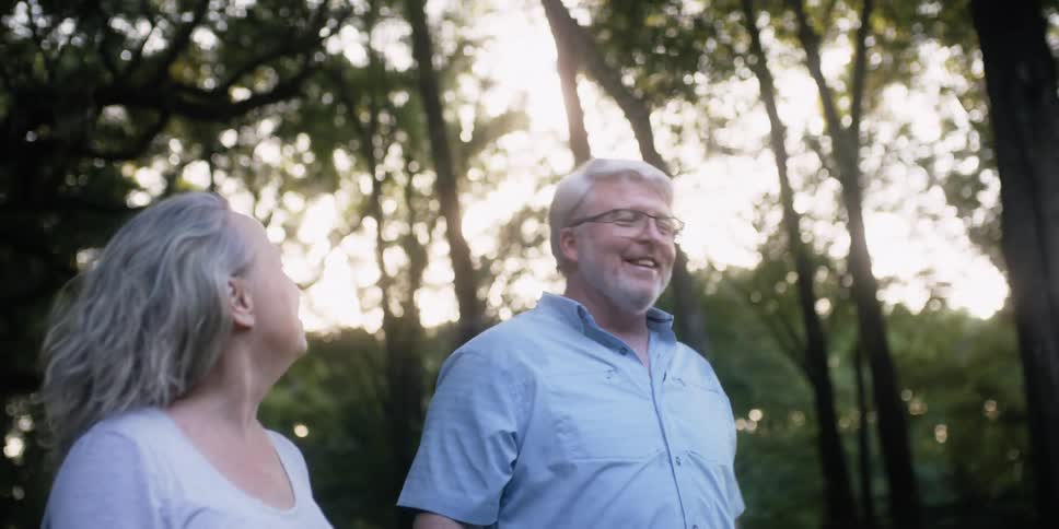 A man and woman look at each other and smile as they walk together near trees