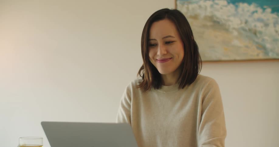 Woman with brown hair and beige sweatshirt uses laptop smiling