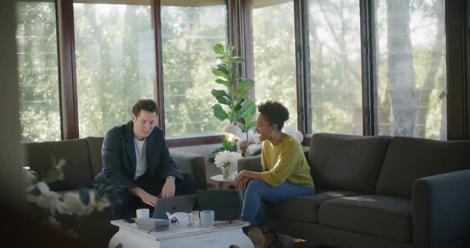 A couple sit on separate couches using their laptop and tablet