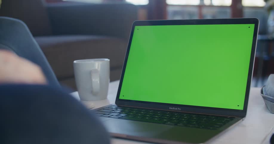 A person sits on a couch at home in front of a laptop with a green screen