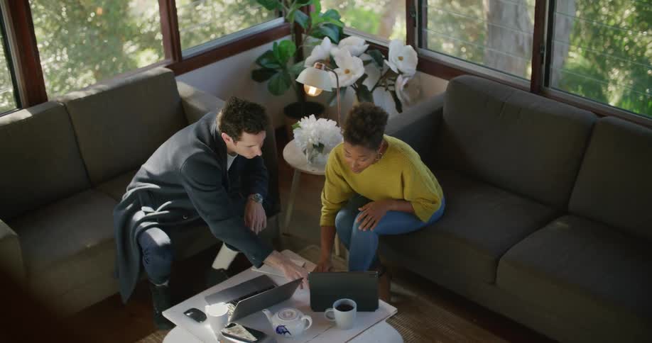 A man and woman sit on couches talking about whats on the two laptops in front of them