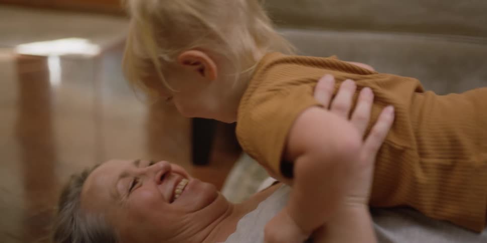 A toddler runs through a living room and into her grandmothers arms as she lies on the floor