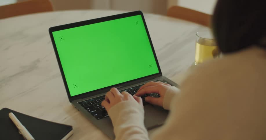 Woman with dark hair and beige sweatshirt sits at table and works on laptop with green screen