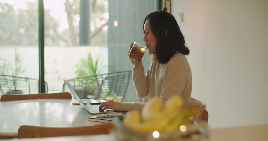 Woman with dark hair and beige sweatshirt drinks a cup of tea while working on laptop