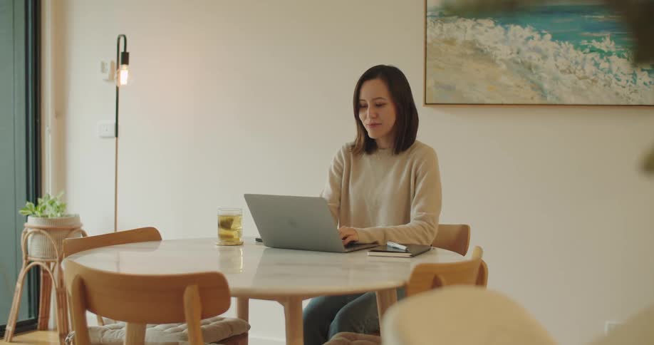 A woman sits at a table at home and types on her laptop