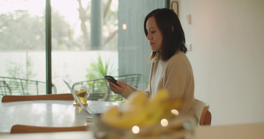 A woman sits at a round dining table typing on a laptop before answering her phone