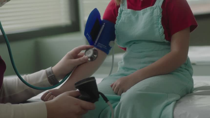 A doctor in a clinic using a stethoscope to listen to a girls pulse with a window and a wall in the background