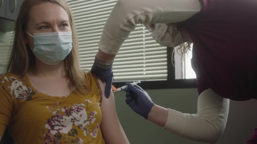A woman with a mask sitting in a clinic and getting an injection from a needle from a nurse
