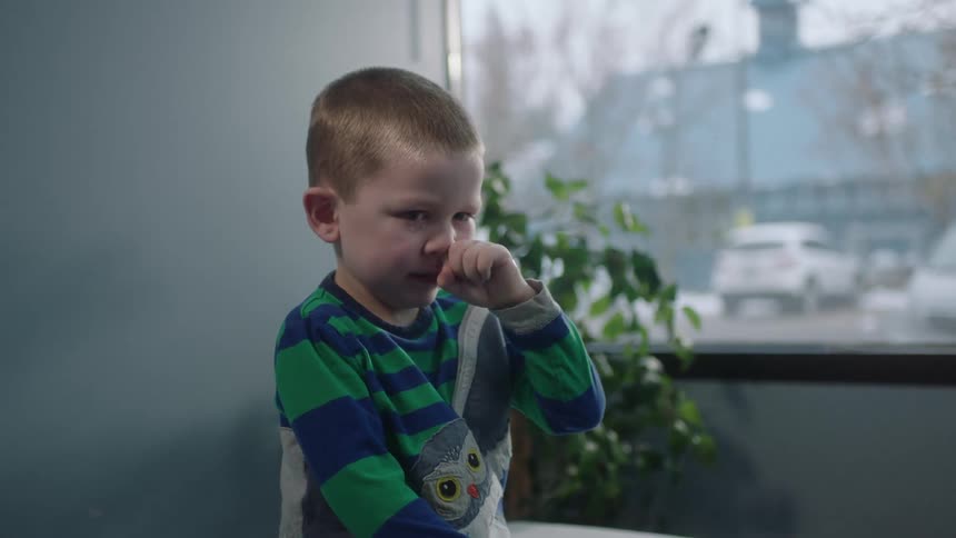 A young boy stands in front of a window and raises his hand to wipe his nose