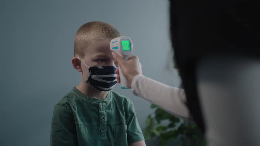 A boy and a nurse in a clinic with the nurse using a thermometer on the boys forehead with a wall and plant in the background