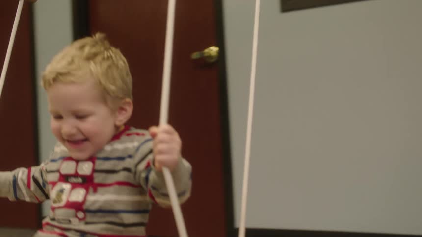 Child swings on wide wooden swing indoor over floor mat