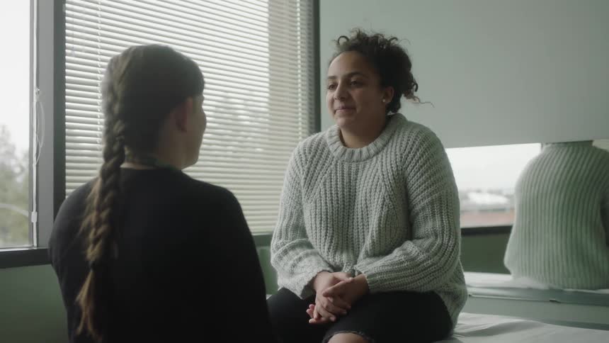 A woman sits on a doctors exam table and talks to a female in front of her