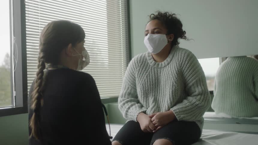 A young female patient sits on an exam table in a doctors office while talking to a female doctor
