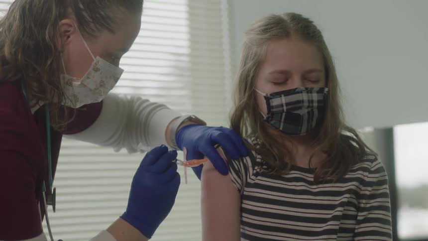 A girl and a nurse in a clinic with the nurse injecting a needle in the girls arm with a window in the background