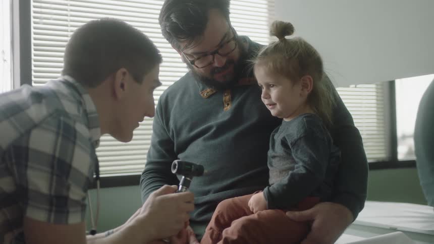 A father holds his toddler daughter on a medical table while a doctor examines her