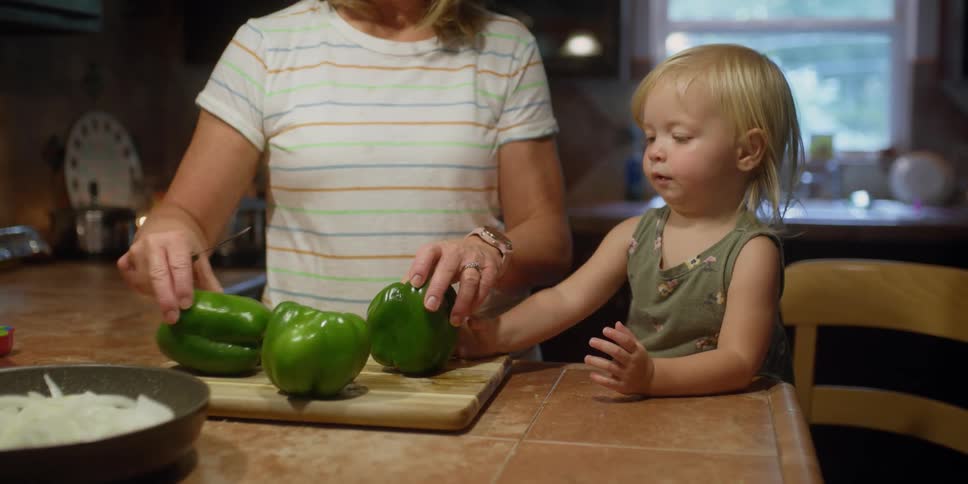 A woman arranges peppers on a cutting board as a young girl standing on a chair watches
