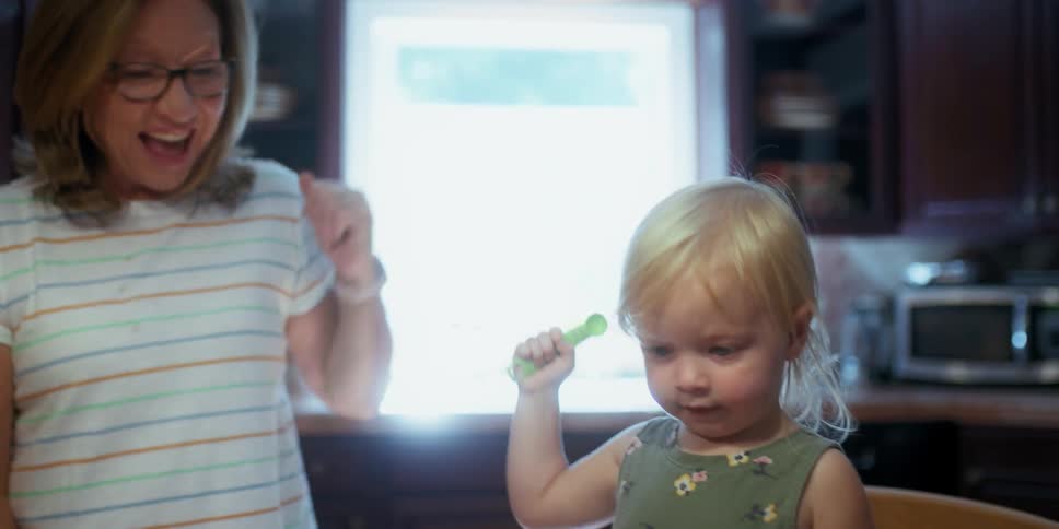 A woman raises her arm and cheers on a little girl holding a spoon in the kitchen