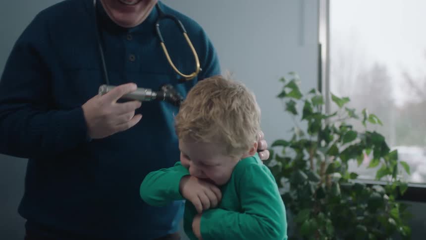 A boy and a doctor in a clinic with the doctor looking into the boys ear with a wall and window in the background