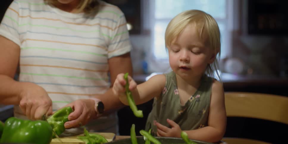 A woman and a girl in a kitchen with a woman cutting a pepper with a knife
