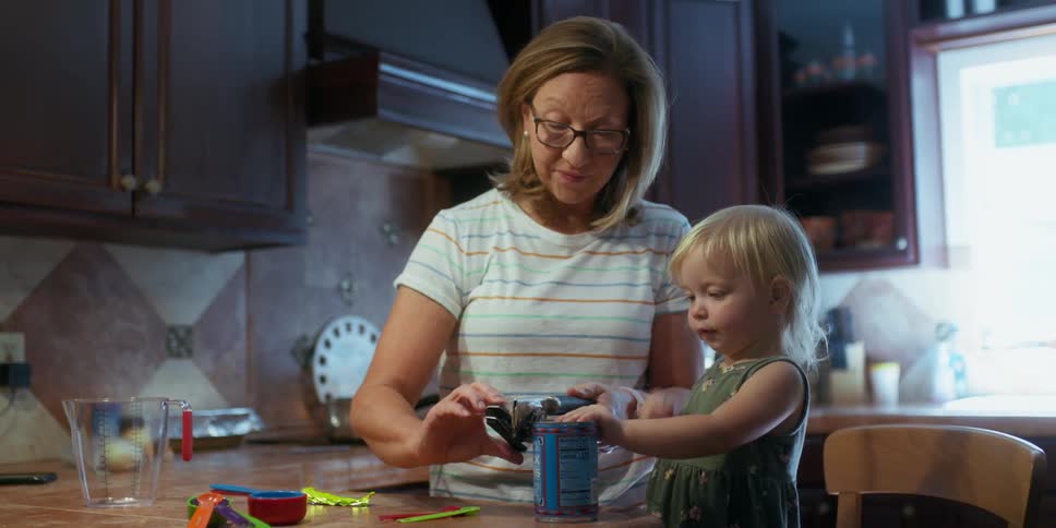 A little girl stands on a chair in a kitchen and helps her grandma open a can
