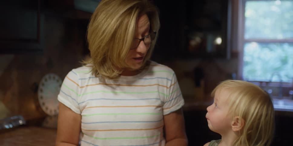 Blonde grandmother with glasses talks to granddaughter putting onion slices in pan in the kitchen