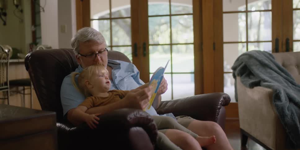 Grandfather with glasses and blue shortsleeved shirt reads story book for his granddaughter sitting in armchair