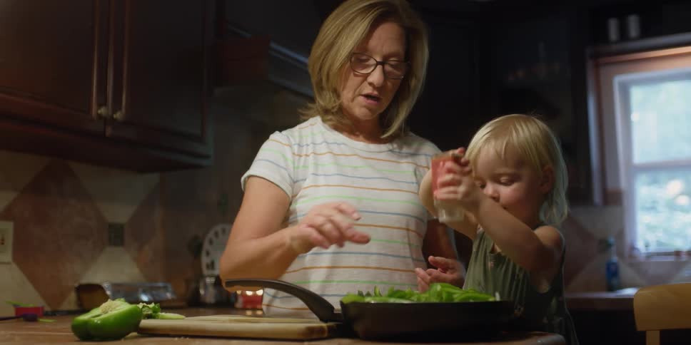 A toddler shakes seasoning in a pan as she helps her mother cook