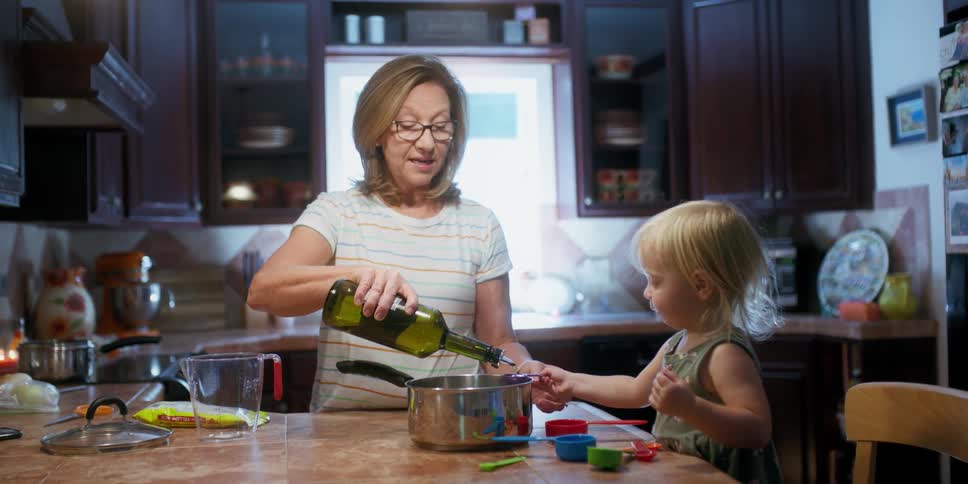 A woman and a girl in a kitchen with the girl pouring olive oil into a pot
