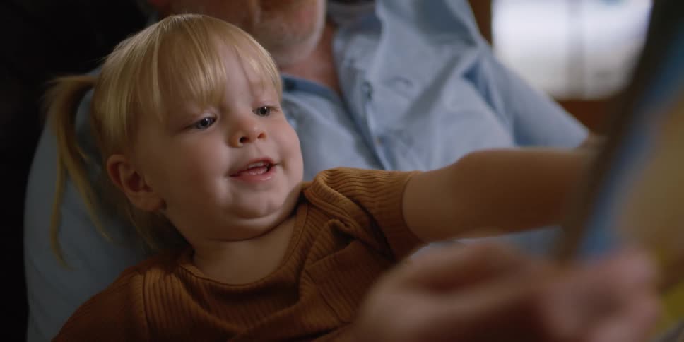 A toddler laughs while sitting in her grandfathers lap while he reads to her