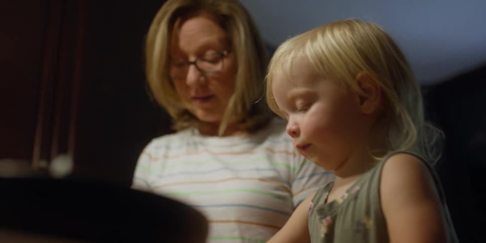 A young toddler places pieces of food in a bowl that a woman gives to them