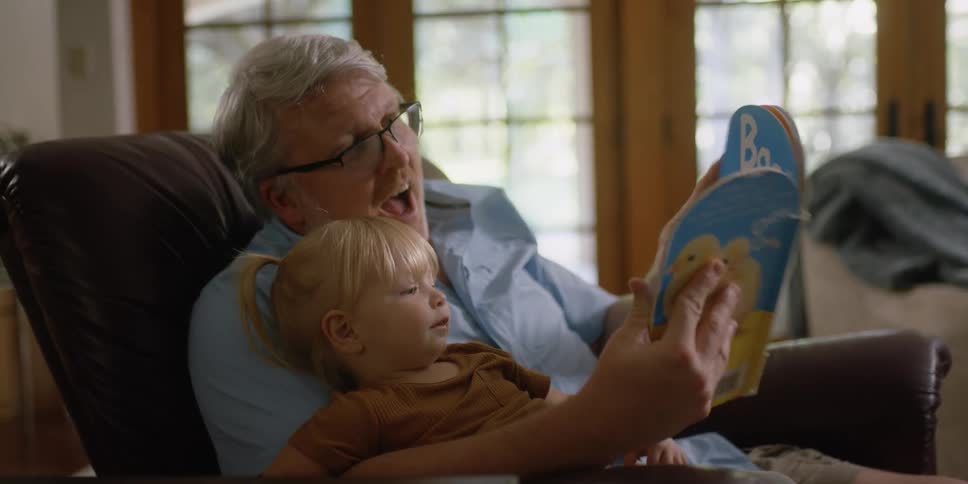 A man sitting in a rocking chair reads a picture book to a young girl sitting in this lap