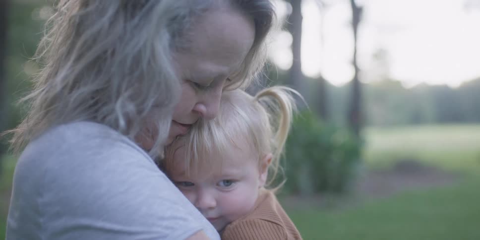 A woman holds and comforts a small girl in her arms while sitting in a field during the day