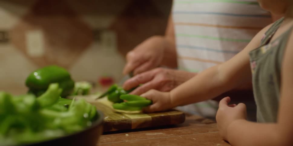 A woman slices peppers with a knife and slides them across the cutting board to a young girl who lifts the slices and places them in a skillet