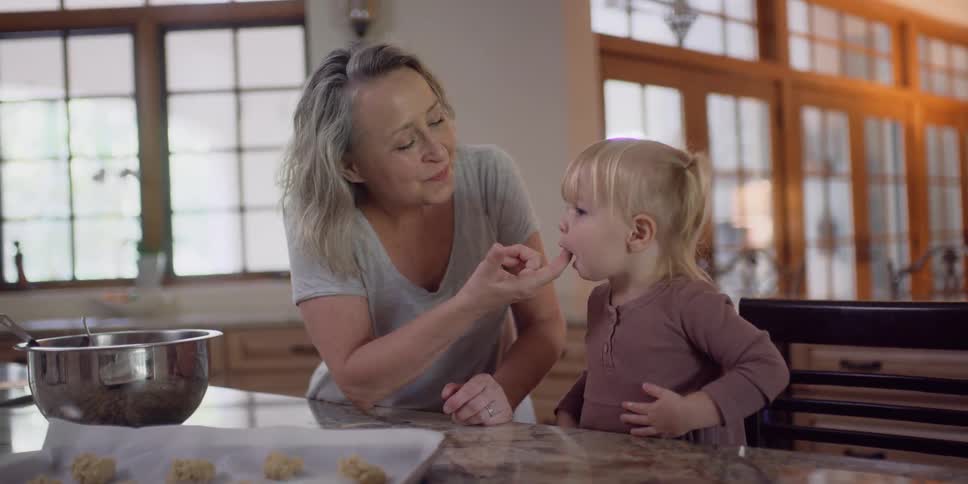 A woman dips her finger in a mixing bowl uses it to feed a young girl a bit of dough