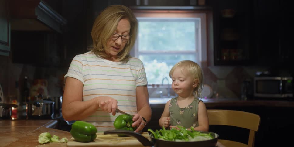 A young toddler stands on a chair and helps a woman cut a fresh bell pepper