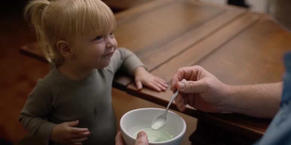 A man feeding a little girl with a spoon and bowl next to a table