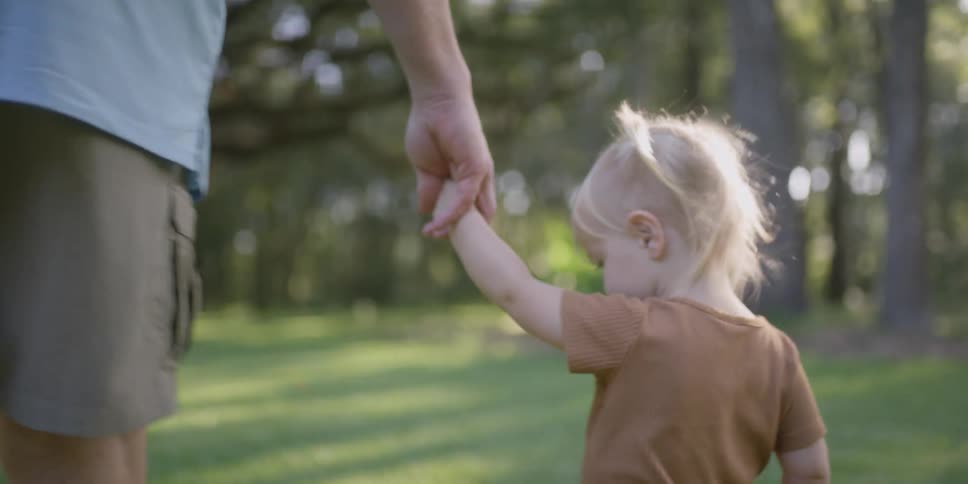 A toddler girl holds her dads hand as they walk through a rural grass yard