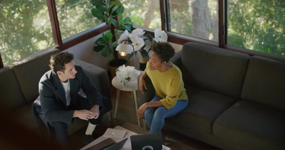 A man and woman sit on couches talking and look to a laptop in front of them