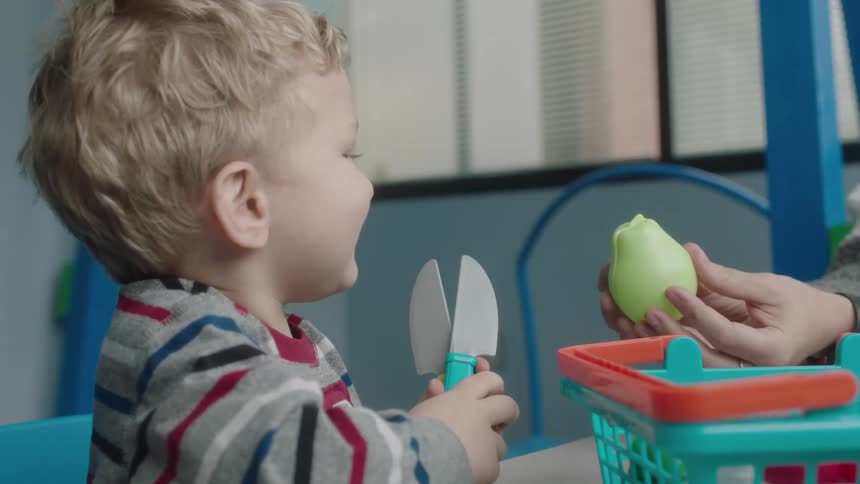 An adult off camera is holding a pear up to a small boy who is holding plastic knives