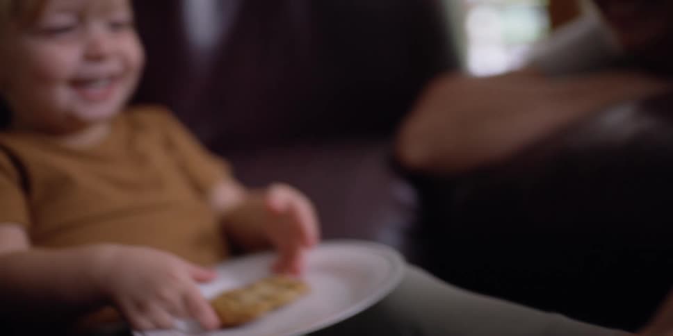A young girl laughs as her father tickles her feet as she sits and eats on a leather couch