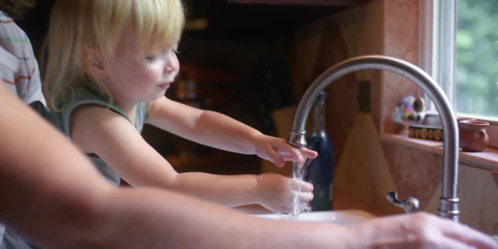 Mother holds daughter helping her reach faucet as they watch their hands in kitchen sink near window