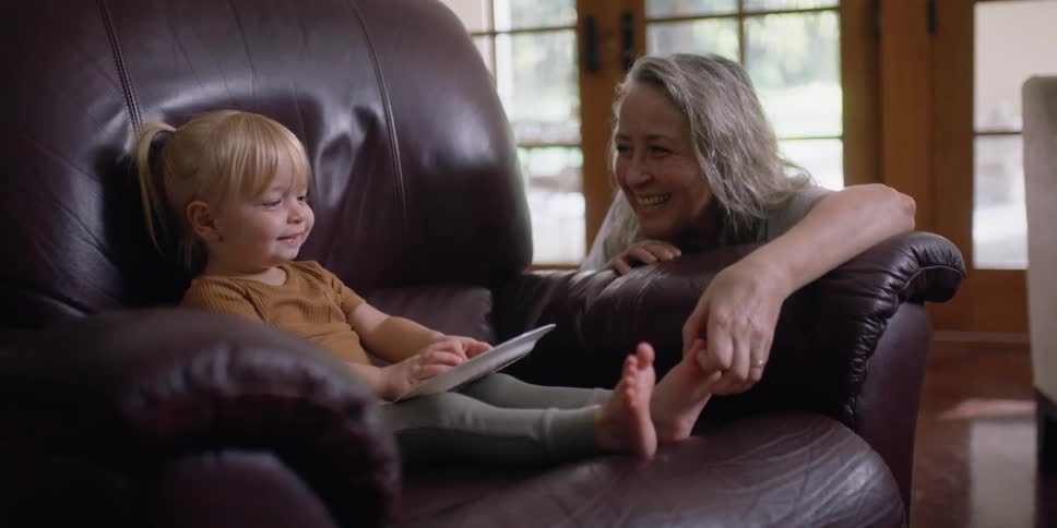 A little girl sits in a chair and eats a cookie as her grandma tickles her toes